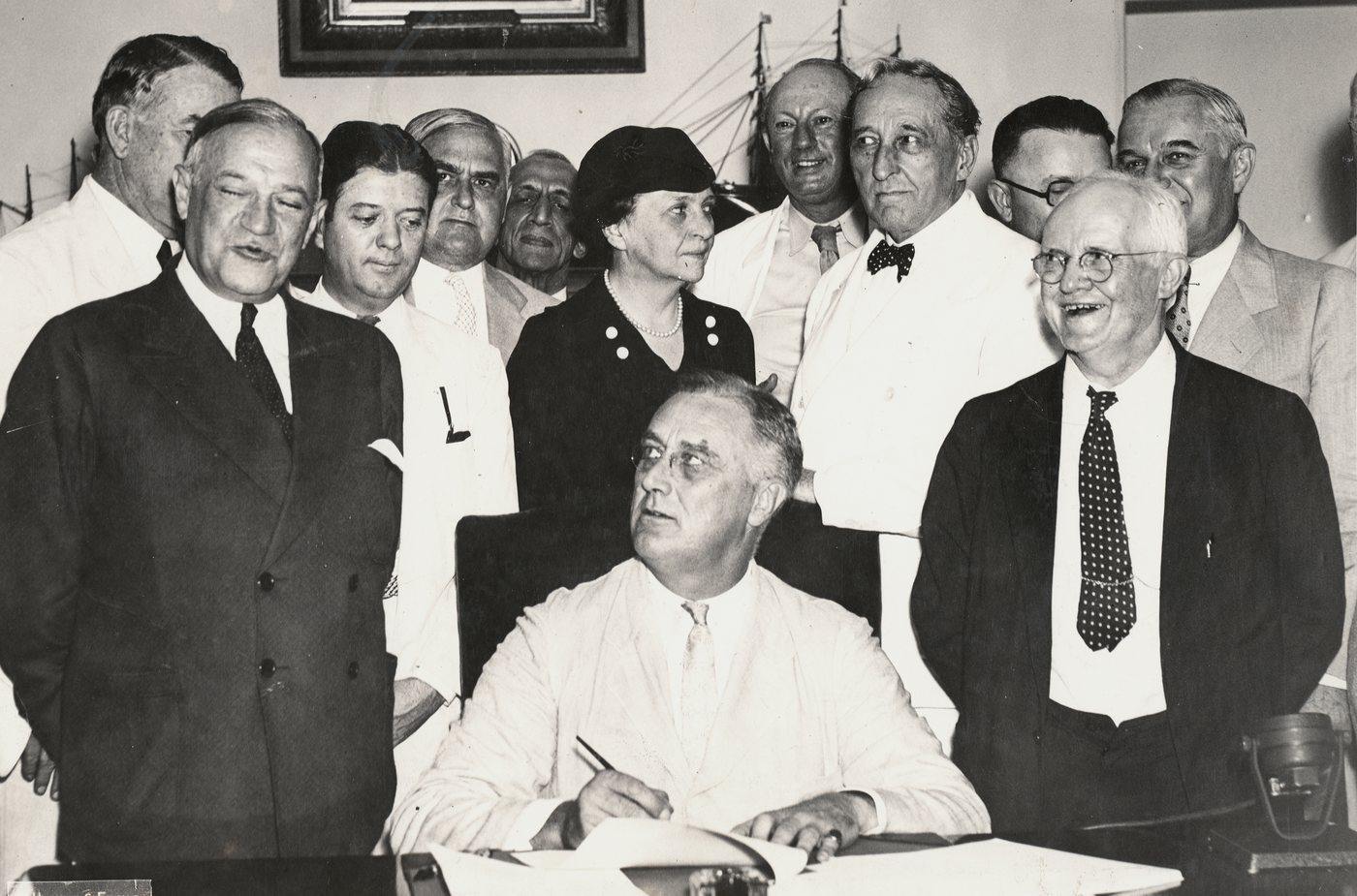 President Roosevelt seated at a desk signing the Social Security Act, officials standing behind him